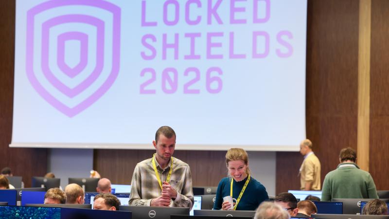 Bildē redzams ekrāns ar uzrakstu "Locked Shields 2026" un fonā smaidīgi cilvēki, kas piedalās šajās mācībās.